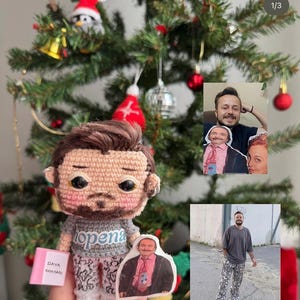 Könnte beinhalten: Eine gehäkelte Puppe mit grauem Oberteil und gemusterter Hose wird vor einem Weihnachtsbaum gehalten. Die Puppe hat einen Bart und ein kleines Bild eines Mannes. Die Puppe hält ein rosa Buch.