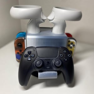 Könnte beinhalten: Ein schwarzer PlayStation 5-Controller liegt auf einem silbernen Ständer vor einer Ladestation für Spielkonsolen. Die Station hält weiße VR-Controller und farbenfrohe Nintendo Switch Joy-Cons. Ein Gaming-Zubehör.