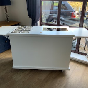 May include: White mobile serving cart with a fold-down counter top and a built-in stainless steel condiment holder. The cart has wheels and is perfect for serving food or drinks.