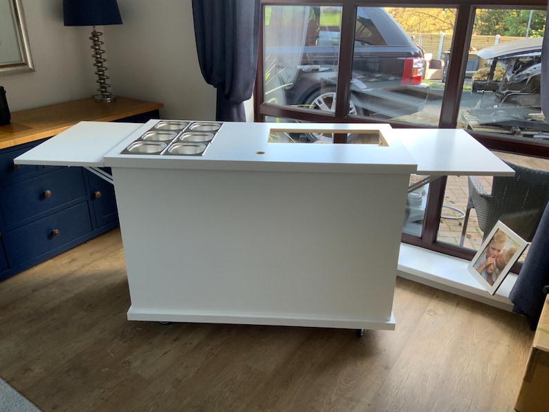 May include: White mobile serving cart with a fold-down counter top and a built-in stainless steel condiment holder. The cart has wheels and is perfect for serving food or drinks.