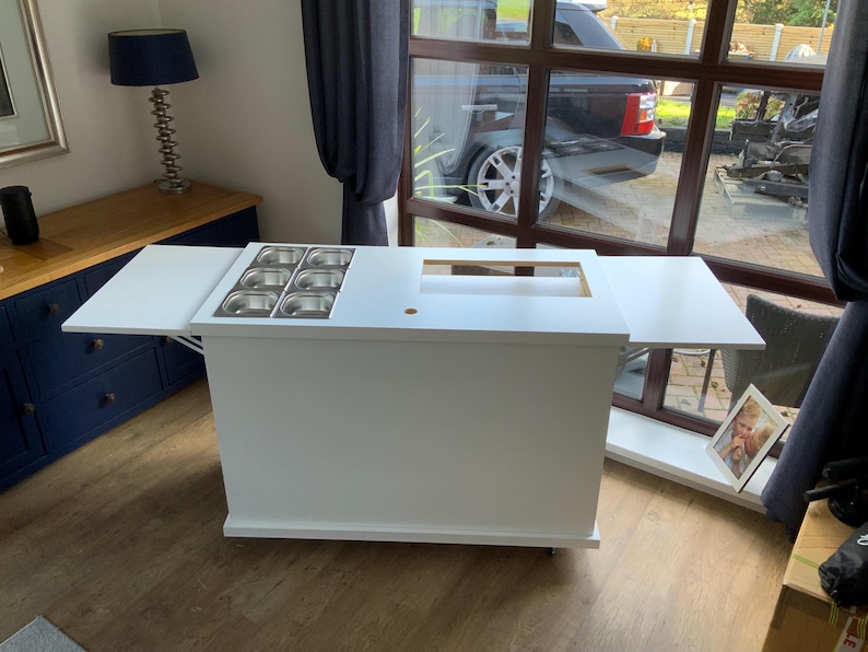 May include: White mobile serving cart with a fold-down side table and a built-in stainless steel condiment holder. The cart has a white countertop with a cut-out for a serving area.