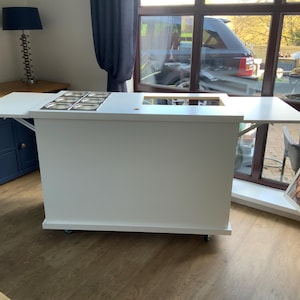 May include: White kitchen island with a drop-down counter top and a built-in stainless steel sink. The island has wheels and is on a wood floor.