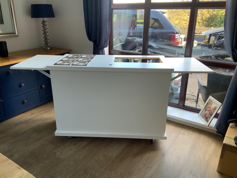 May include: White kitchen island with a drop-down counter top and a built-in stainless steel sink. The island has wheels and is on a wood floor.