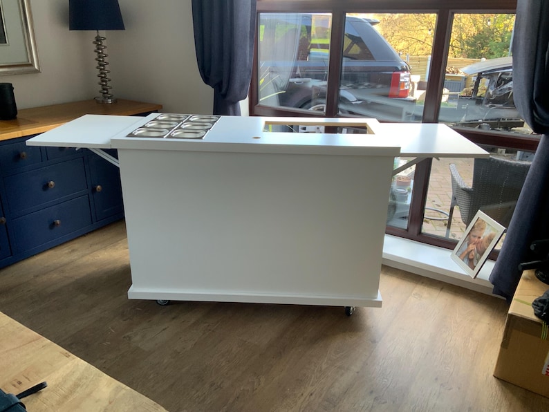 May include: A white mobile bar cart with a fold-down countertop and a built-in stainless steel ice cream topping station. The cart has wheels and is on a wood floor.