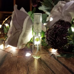 May include: A clear glass bottle with a silver spray nozzle filled with a light yellow liquid. The bottle is surrounded by white flowers and green leaves.