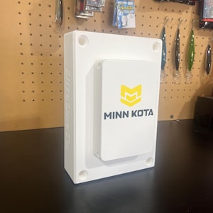 May include: A white rectangular device with the text "MINN KOTA" and a yellow logo. The device is mounted on a larger white rectangular base. Fishing lures and hooks are visible in the background.