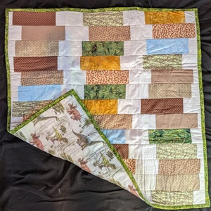 May include: A white baby quilt with a dinosaur print on the back. The front of the quilt is made of various fabric squares in shades of brown, green, yellow, and blue. The quilt is edged with a green trim.