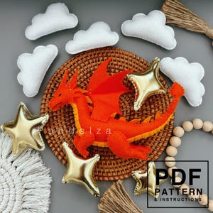 Op de afbeelding: Een feloranje vilt draak met gele accenten ligt op een gevlochten mand, omringd door witte vilten wolken en gouden sterornamenten. De afbeelding bevat de tekst "PDF PATTERN & INSTRUCTIONS."