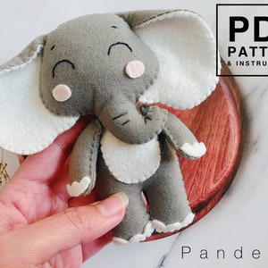 May include: A gray felt elephant plush toy with white felt accents. The elephant has a stitched smile and pink felt cheeks. The toy is being held in a hand. The text "PDF PATTERN & INSTRUCTIONS" is in the upper right corner of the image. The text "Pandelza" is in the lower right corner of the image.