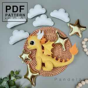 Op de afbeelding: Een gele vilt draak met oranje vleugels en een staart, zittend op een geweven rieten dienblad met gouden vilt sterren en witte vilt wolken. De tekst "PDF PATTERN & INSTRUCTIONS" staat in een zwarte doos bovenaan de afbeelding.