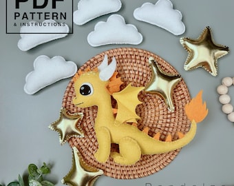 May include: A yellow felt dragon with orange wings and a tail, sitting on a woven wicker tray with gold felt stars and white felt clouds. The text "PDF PATTERN & INSTRUCTIONS" is in a black box at the top of the image.