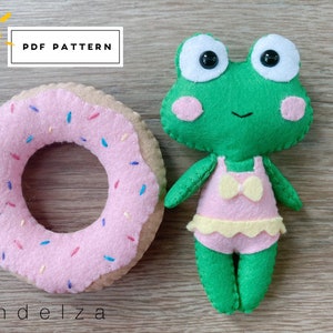May include: A green felt frog wearing a pink swimsuit with a yellow bow. The frog is holding a pink and white donut with sprinkles.