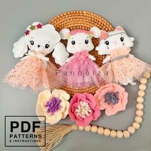 Op de afbeelding: Drie vilten dierenpoppen gekleed in roze en witte tule rokjes. De poppen zijn een witte poedel, een witte muis en een witte kat. De poppen zijn gerangschikt op een geweven mand met een houten kralensnoer en vilten bloemen.