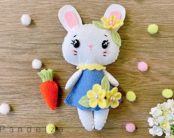 Può includere: Una bambola di coniglio di feltro bianco che indossa un vestito blu con fiori gialli e una perla. Il coniglio ha occhi neri e un naso rosa. La bambola tiene una carota di feltro nella sua mano. La bambola è su una superficie di legno con palline di feltro bianche e verdi.