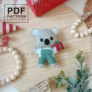 Puede incluir: Un koala de fieltro gris que lleva una camisa blanca y un mono verde sostiene una caja de regalo roja. El koala está sentado sobre una superficie de madera con una guirnalda de cuentas de madera y una pequeña planta verde.