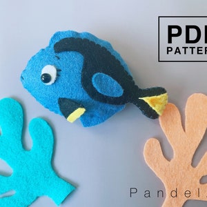 May include: A blue and yellow felt fish with a black eye and a black and yellow tail fin. The fish is surrounded by two felt coral pieces, one blue and one orange. The text "PDF PATTERN" is in a black box in the upper right corner of the image. The text "P and elza" is in the lower right corner of the image.