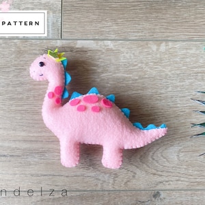 May include: A pink felt dinosaur with blue and pink accents, wearing a gold crown. The dinosaur has a smiling face and is on a wooden surface. The text "PDF PATTERN" is in a white box in the upper left corner.