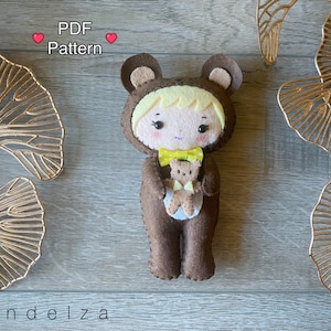 May include: A brown felt bear plush toy wearing a yellow bow tie and holding a smaller brown felt bear plush toy. The larger bear has a yellow and white face with black eyes and a pink nose. The text "PDF Pattern" is above the bear.