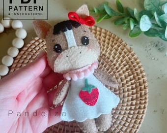 May include: A felt toy horse wearing a white dress with a red strawberry design and a pink bow. The horse has brown fur, a white stripe down its face, and black eyes.