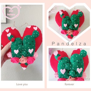 Puede incluir: Una decoración de pared en forma de corazón de fieltro con dos figuras de cactus verdes abrazándose. Las figuras de cactus tienen corazones rosas y ojos blancos. El corazón está rodeado de flores de fieltro rosas y blancas. El texto "Love you" está escrito debajo del corazón. El texto "PDF Pattern" está escrito en un corazón rosa en la parte superior de la imagen.