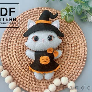 May include: A grey felt cat wearing a black witch hat with an orange band and a black dress with an orange pumpkin on the front. The cat is sitting on a woven brown wicker surface.
