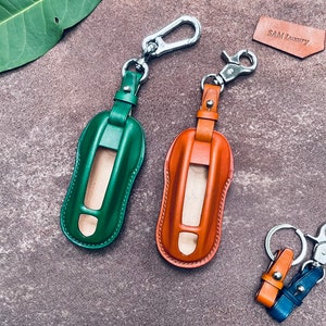 May include: Two leather car key fob covers in green and orange, each featuring a silver metal swivel snap hook. A smaller orange and blue leather keychain with a silver split ring and snap hook is included, alongside a small leather tag labelled SAM Luxury.
