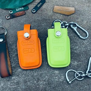 Peut inclure: Divers porte-clés et porte-clés en cuir de différentes couleurs. Les porte-clés sont orange et vert citron, avec des accessoires argentés. Plusieurs petites étiquettes en cuir gravées sont également présentes, avec des noms comme "Alicia" et "Lowe".