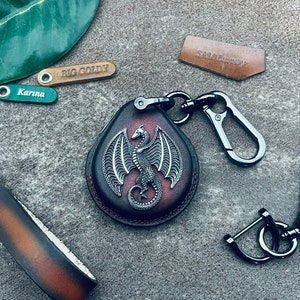 Könnte beinhalten: Ein Leder-Schlüsselanhänger mit einem silbernen Drachen-Design. Der Schlüsselanhänger ist rot und braun mit einem silbernen Metallverschluss und Ring.