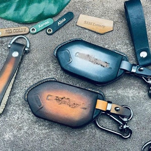 May include: Two leather keychains with key fobs. One keychain is brown with a gold accent and the other is black with a silver accent. Both keychains have a metal clasp and a leather strap. The key fobs are both made of leather and have a textured surface. The key fobs are both shaped like a car key. The keychains are labeled with the names "BIG GOLDY", "Karina", "Alicia", "SAM Luxury".