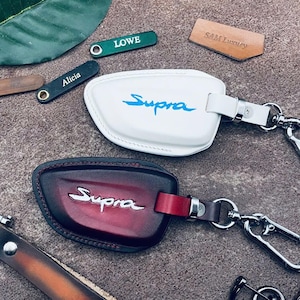 May include: Two leather keychains with metal rings and clips. Both keychains have the word "Supra" embossed on them. One keychain is white leather with blue lettering and the other is red leather with silver lettering.
