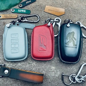 May include: Four different keychains with leather covers. The keychains are silver, red, black, and brown. The leather covers have different designs, including a car logo, a Sasquatch, and a series of buttons. The keychains are on a gray surface.