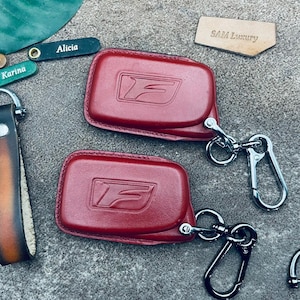 May include: Two red leather keychains with a silver F logo and a silver carabiner clip. The keychains are on a gray surface.