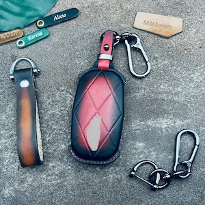 May include: A collection of leather keychains and key fobs. The key fob is black and red with a diamond pattern. Other keychains are brown and green with names like "Alicia" and "Karina" engraved. A tag reads "SAM Luxury".