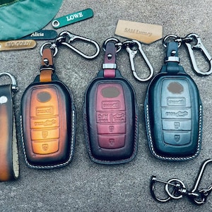 May include: Three leather key fobs with different colored designs. The key fobs are brown, red, and gray. Each key fob has a metal ring and a metal hook. The key fobs are labeled "BIG GOLDY", "LOWE", and "SAM Luxury".