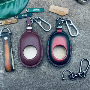 May include: Two leather keychains with a key ring and a small pocket for a key fob. The keychains are burgundy and black with a red accent. The keychains are on a gray surface with other keychains and tags.