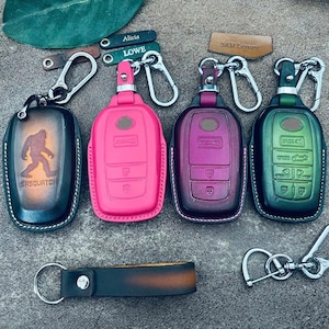 Puede incluir: Cuatro fundas de cuero para llaves de coche en negro con un gráfico de Sasquatch, rosa brillante, morado oscuro y verde bosque, cada una con un mosquetón de metal plateado. Incluye etiquetas de cuero con nombres como Alicia y LOWE, y un llavero negro.