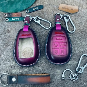May include: Two leather car key fobs in a deep burgundy color with white stitching. One fob has a keyhole cutout, the other has buttons labeled "Panic", and car symbols. Keychains and small tags with names are also present.