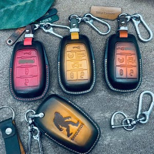 May include: Three leather car key fob covers in red, yellow, and brown gradients with silver metal clips, plus one brown leather key fob cover featuring an embossed Sasquatch silhouette and the text SASQUATCH, all with visible white stitching.