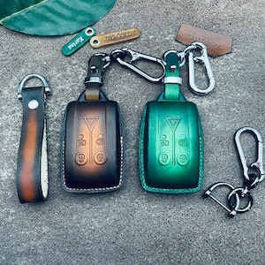 May include: Two leather keychains with key fobs. One keychain is brown with a black fob and the other is green with a silver fob. Both fobs have a triangle and three buttons. The keychains have metal rings and leather straps.