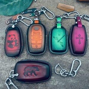 May include: Five leather key fob covers with metal carabiner clips. One is red with a panda, one is brown, one is green with a dragonfly and letter M, one is magenta with a cross, and one is dark red with a bear silhouette labeled mama.