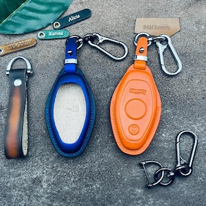 Könnte beinhalten: Zwei Leder-Schlüsselanhänger mit Metallschlüsselringen. Ein Schlüsselanhänger ist blau und der andere orange. Beide Schlüsselanhänger haben ein Autologo. Die Schlüsselanhänger liegen auf einer grauen Oberfläche.