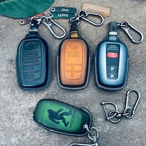 May include: Four leather car key fob covers with metal carabiner clips. Three covers are upright in black, tan, and grey, featuring embossed button icons. One green cover lies horizontally, embossed with a Sasquatch figure and the text SASQUATCH.