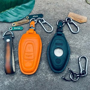Könnte beinhalten: Eine Auswahl an Leder-Schlüsselanhängern in Orange, Dunkelgrün und Braun. Der orangefarbene Schlüsselanhänger hat geprägte Autoschlüsselsymbole. Zusätzliche Schlüsselanhänger sind mit "Alicia", "Big Goldy" und "Karina" beschriftet. Ein Etikett lautet "SAM Luxury".