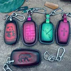 May include: A collection of leather car key fob covers in various colours including red, purple, and green, featuring embossed designs like a panda, a cross, a dragonfly, and a bear with the text mama, all attached to silver metal key rings.