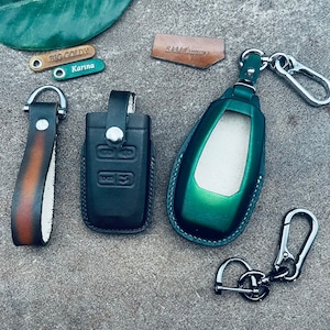 Peut inclure: Trois porte-clés en cuir avec des cuirs et des anneaux de clés métalliques de différentes couleurs. Un porte-clés a un porte-clés en cuir noir avec un bouton argenté et le texte "BIG GOLDY" sur une étiquette en cuir marron. Un autre porte-clés a un porte-clés en cuir vert avec un anneau de clé argenté. Le troisième porte-clés a une sangle en cuir marron avec un insert en cuir blanc et un anneau de clé argenté.