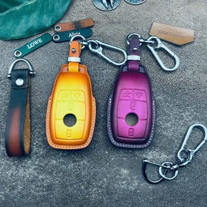 Peut inclure: Deux porte-clés en cuir avec un design dégradé jaune et violet. Les porte-clés ont un anneau en métal argenté et un crochet en métal argenté. Les porte-clés sont sur une surface grise. Le texte "SAM Leather" est sur une étiquette en cuir marron.