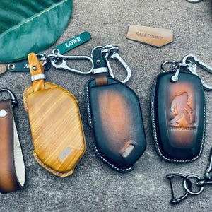 May include: Three leather key fobs with metal hardware. One is light brown with a wood grain pattern, one is a dark brown to black gradient, and one is black with a brown centre and a debossed Bigfoot silhouette with the text "SASQUATCH".