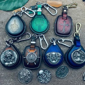 May include: A collection of six leather keychains with different designs. The keychains feature a skull, a cross, an anchor, a skeleton, a tree, and a hand. The keychains are all made of brown leather and have silver accents.