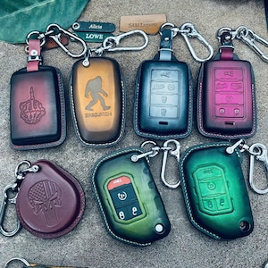 Peut inclure: Divers étuis de clés en cuir de différentes couleurs, dont bordeaux, vert et noir. Certains étuis présentent des motifs en relief tels qu'un majeur, un Sasquatch et un crâne. Chaque étui est muni d'un porte-clés en métal.