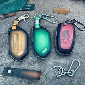 Könnte beinhalten: Drei Schlüsselanhänger-Etuis aus Leder mit Metallclips. Eines ist braun mit geprägter amerikanischer Flagge, eines ist einfarbig grün und eines ist rot mit einer schwarzen Sasquatch-Silhouette und dem Text "SASQUATCH". Ein Lederband und Clips liegen bei.